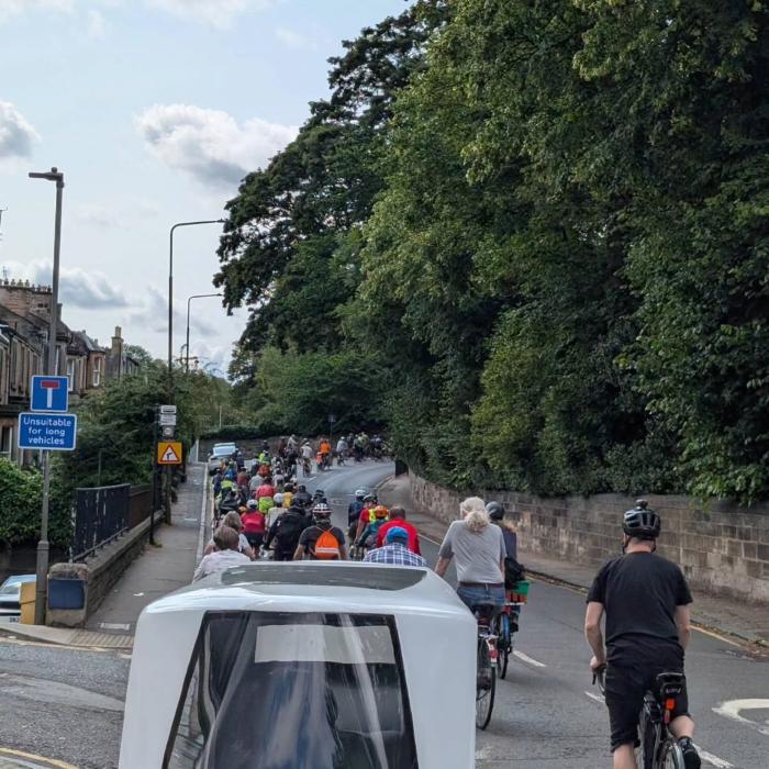 A lane full of cyclist on a two lane wide curved road in West Edinburgh. Blue sky, full trees along the right side of the road and 2 story Victorian residential buildings on the left.